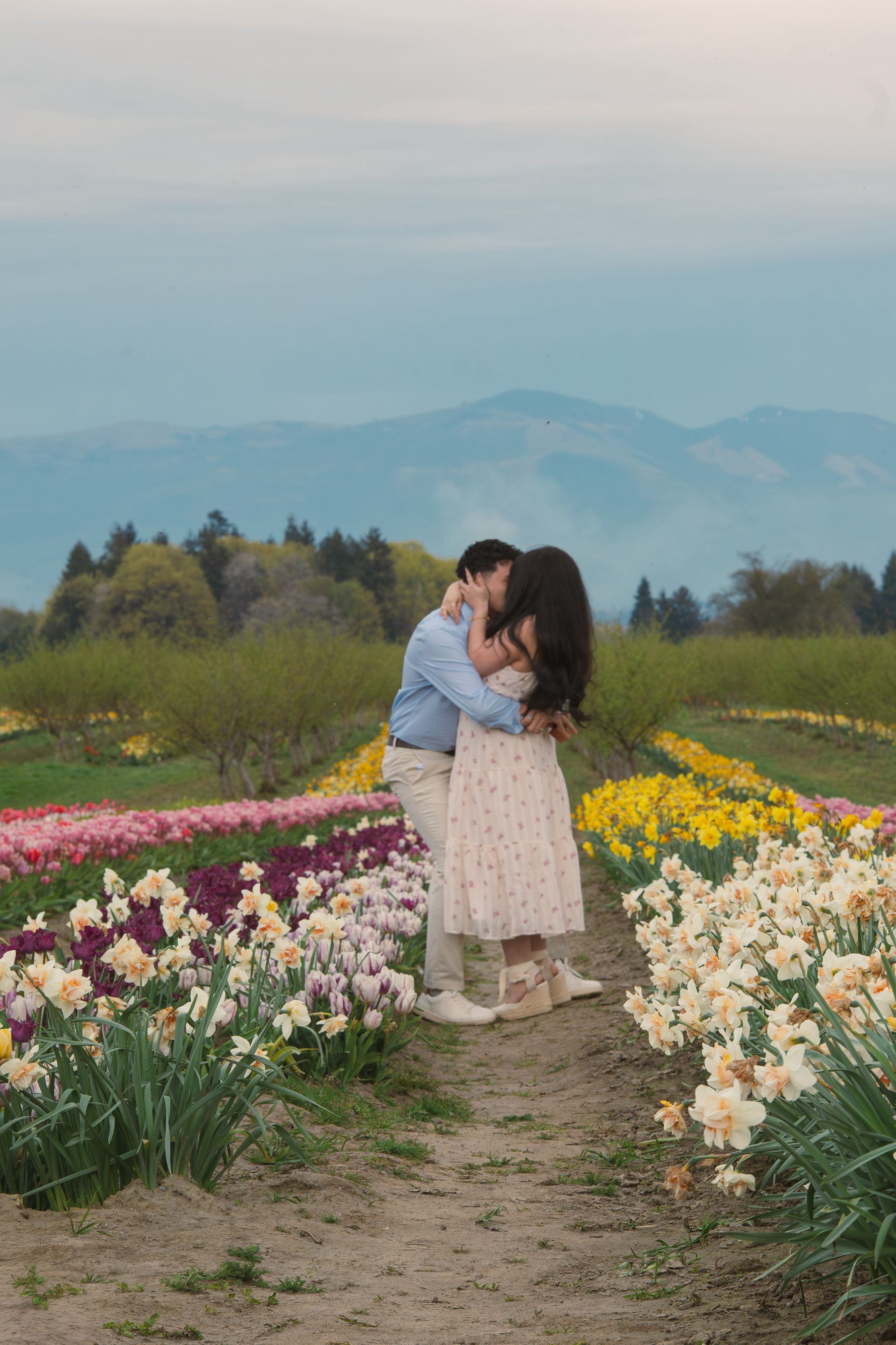 A man and woman kiss between rows of flowers with trees and mountains in the background