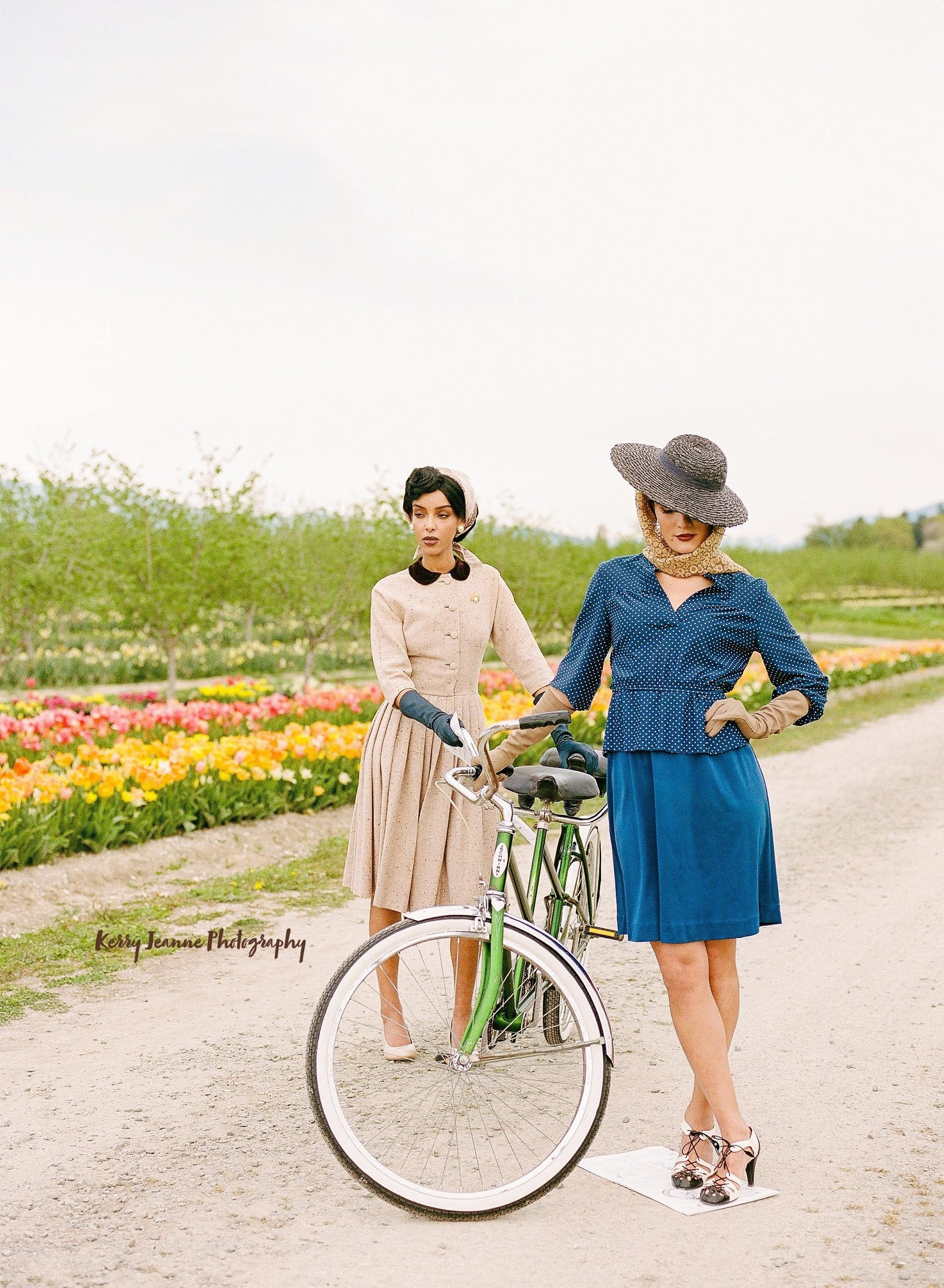 Two women in period clothing pose with a bike in front of rows of tulips and orchard trees.